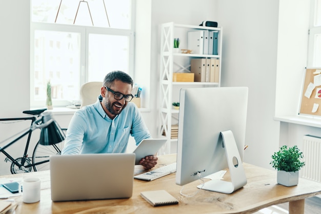 Bell'uomo di mezza età in abbigliamento casual intelligente che utilizza tablet digitale e sorride mentre è seduto in ufficio