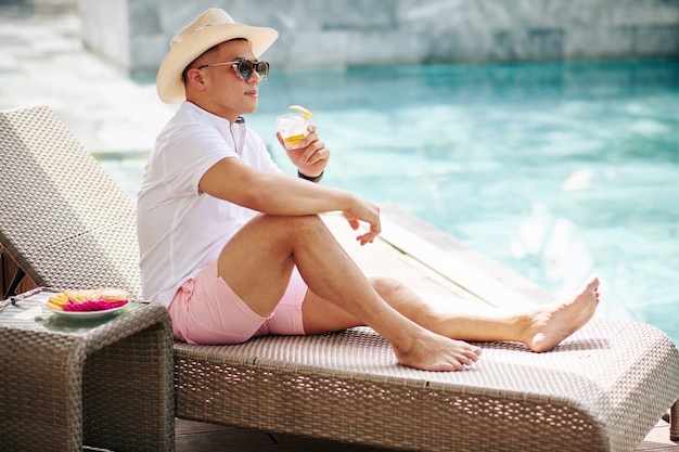 Bell'uomo elegante in cappello e occhiali da sole seduto sulla chaise-longue in piscina e bere gin tonic con buccia d'arancia