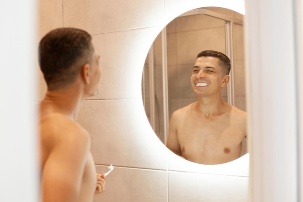 Bell'uomo estremamente felice con il torso nudo in piedi davanti allo specchio in bagno, lavandosi i denti, guardando i denti con il dentifricio e sorridendo.