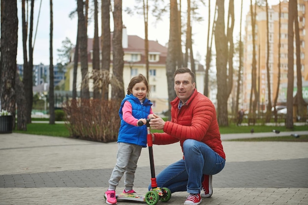 Bell'uomo europeo di mezza età felice padre premuroso e la sua amata figlia su uno scooter da calcio che trascorrono del tempo insieme in un parco urbano