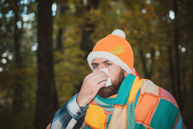 Bell'uomo hipster con la barba avvolta in una coperta multicolore, con cappello caldo, naso che cola. Concetto di resistenza alle malattie stagionali, raffreddori, prevenzione naso che cola, allergia.