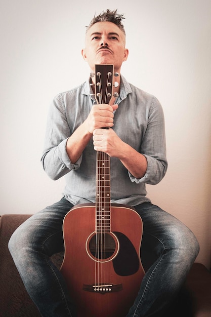 Bell'uomo in abiti casual è in posa con la sua chitarra acustica Ragazzo con la chitarra seduto sul divano in soggiorno Uomo attraente che tiene la chitarra nelle sue mani Amante della musica Lifestyle Hobby concept