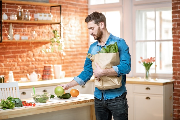 Bell'uomo in camicia blu che disimballa cibo sano dalla borsa della spesa in cucina