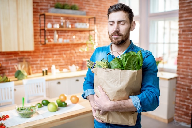 Bell'uomo in camicia blu in piedi con la borsa della spesa piena di cibo verde sano in cucina