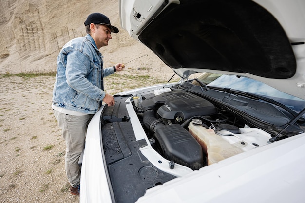 Bell'uomo in giacca di jeans e berretto è in piedi vicino alla sua muscle car bianca con il cofano apribile per controllare il livello dell'olio del motore