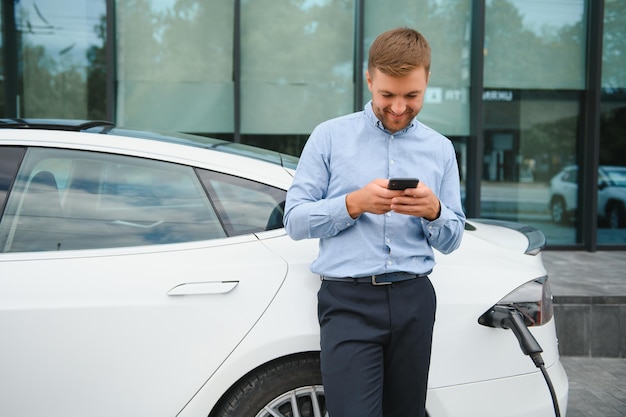 Bell'uomo in giacca e cravatta che naviga in Internet su uno smartphone moderno mentre aspetta la ricarica dell'auto elettrica