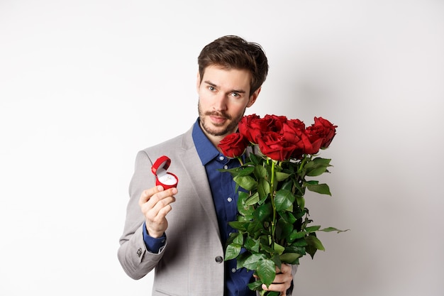 Bell'uomo in tuta, mostrando l'anello di fidanzamento e guardando romantico la fotocamera, in piedi con rose rosse su sfondo bianco. Il giorno di San Valentino e il concetto di amore.