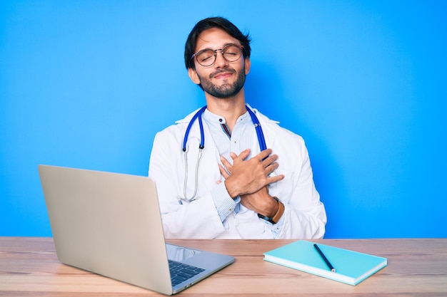 Bell'uomo ispanico che indossa l'uniforme del medico che lavora in clinica sorridendo con le mani sul petto, gli occhi chiusi con un gesto grato sul viso. concetto di salute.