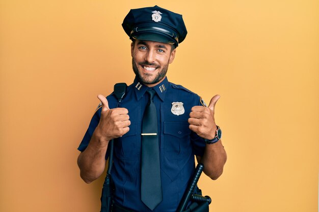 Bell'uomo ispanico che indossa l'uniforme della polizia, segno di successo, facendo un gesto positivo con la mano pollice in alto sorridente e felice espressione allegra e gesto del vincitore