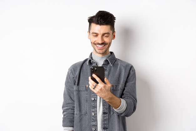 Bell'uomo leggendo il messaggio sul telefono e sorridente. Ragazzo che tiene smartphone e guardando lo schermo, in piedi su sfondo bianco.