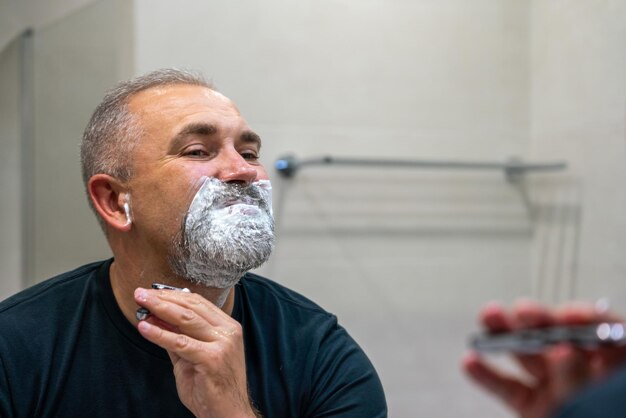 Bell'uomo maturo con i capelli grigi che cerca di tagliarsi la barba da solo in un bagno