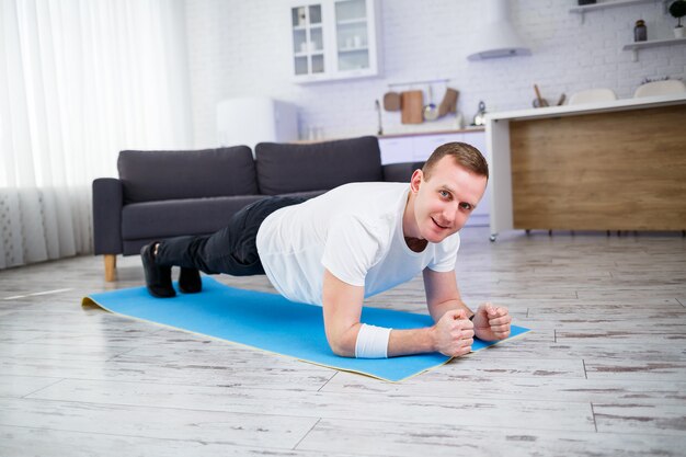 Bell'uomo muscoloso in una t-shirt che fa esercizi di plancia funzionali sul pavimento a casa. Forma fisica a casa. Uno stile di vita sano.