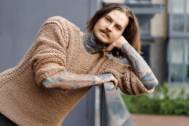 bell'uomo sexy tatuato con capelli lunghi e baffi in un maglione lavorato a maglia. Ritratto al di fuori del primo piano.