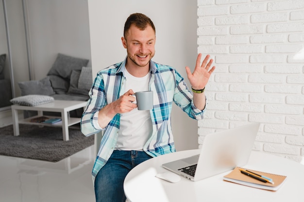 Bell'uomo sorridente in camicia seduto in cucina a casa al tavolo che lavora online sul laptop da casa