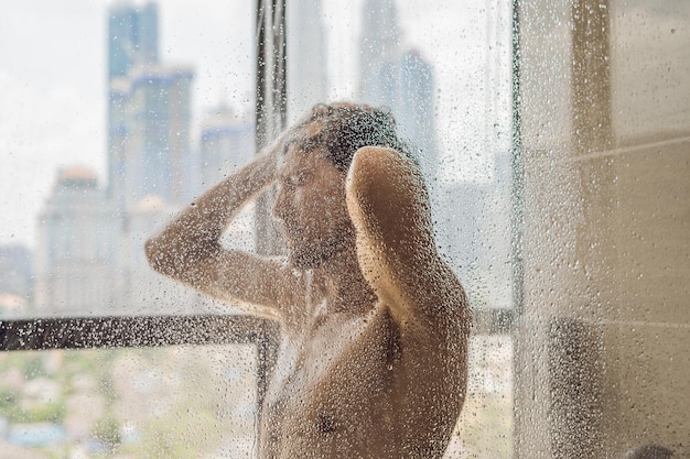 Bell'uomo sotto la doccia dietro un vetro con gocce sullo sfondo di una finestra con vista panoramica della città