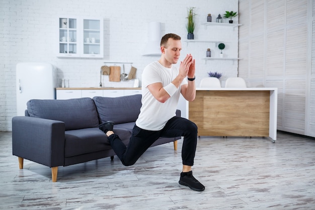 Bell'uomo sportivo con una maglietta che fa squat a casa nel suo appartamento. Forma fisica a casa. Uno stile di vita sano