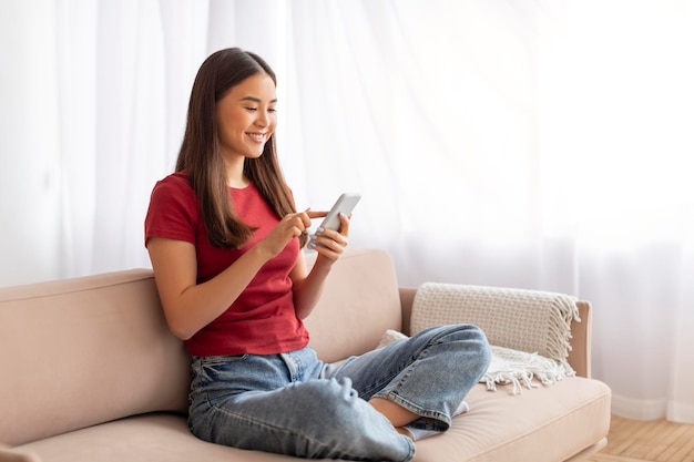 Bella app sorridente donna asiatica che usa il cellulare a casa