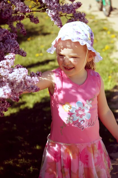 Bella bambina bionda con panama in testa che gioca in giardino primaverile con cespuglio fiorito di lillà viola