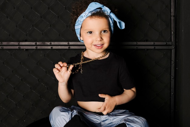 Bella bambina carina con capelli afro e una bandana blu sulla fronte seduta su un barile di metallo su sfondo nero di recinzione metallica