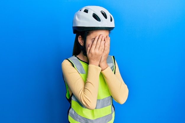 Bella bambina castana che indossa casco da bici e giubbotto riflettente con espressione triste che copre il viso con le mani mentre piange il concetto di depressione