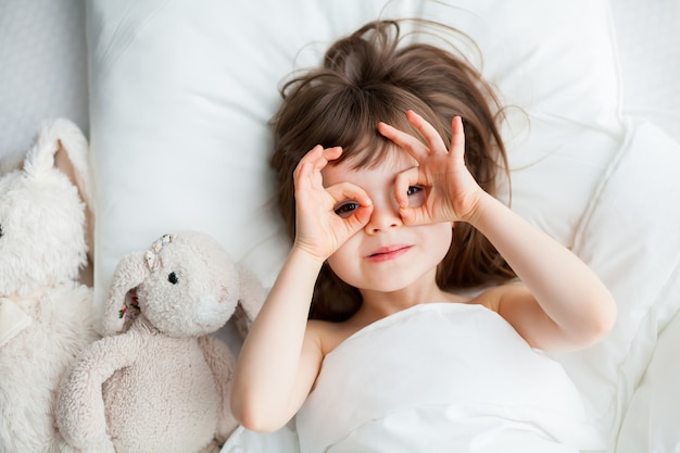 Bella bambina che gioca sciocco sdraiato in un letto bianco con giocattoli di coniglio che si trovano vicino. Vista dall'alto.