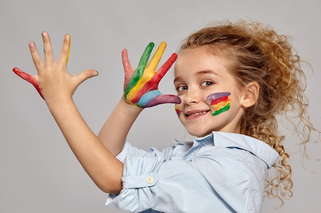 Bella bambina che ha una spazzola tra i suoi capelli biondi ricci chic, indossa una camicia blu e una t-shirt bianca. Sta facendo facce con braccia e guance dipinte, sorridendo su uno sfondo grigio.