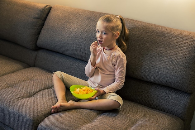 Bella bambina che mangia cibo malsano mentre guarda la tv sul divano