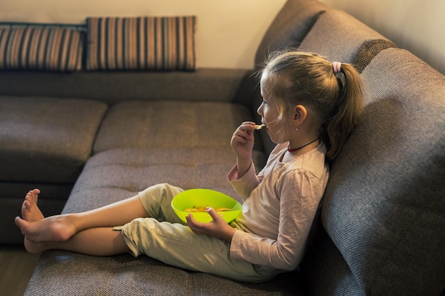 Bella bambina che mangia cibo malsano mentre guarda la tv sul divano