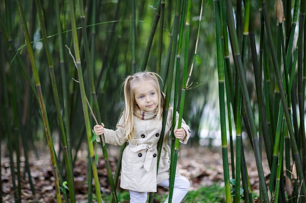 Bella bambina con capelli biondi che stanno in una foresta di bambù e che scuotono bambù