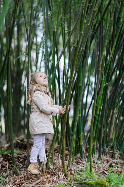 Bella bambina con capelli biondi che stanno in una foresta di bambù e che scuotono bambù