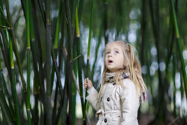 Bella bambina con capelli biondi che stanno in una foresta di bambù e che scuotono bambù