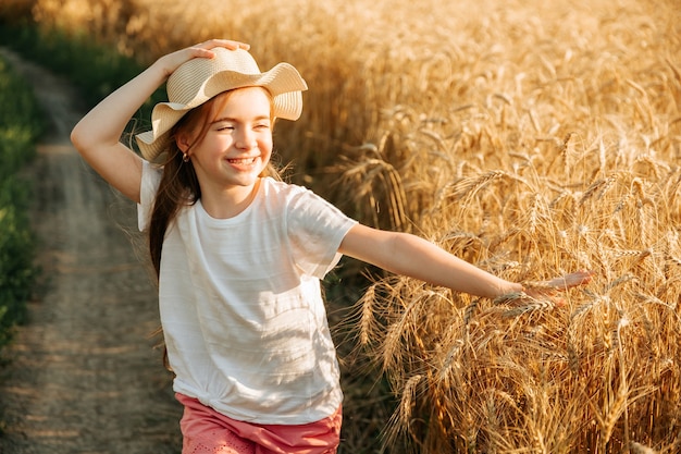 Bella bambina con cappello in testa e sorriso attraente corre sulla strada di campagna vicino al campo...
