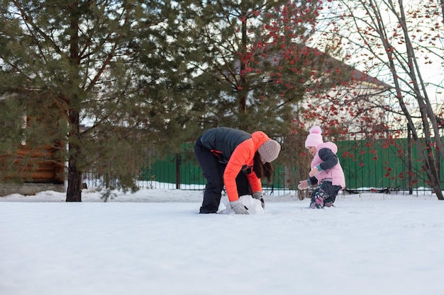 Bella bambina con la madre che rotola palle di neve per costruire pupazzo di neve sul cortile del cottage in serata con sorbo e abeti sullo sfondo Genitori che trascorrono del tempo con i bambini