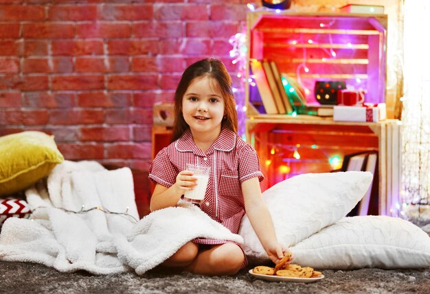 Bella bambina con un bicchiere di latte e gustosi biscotti nella stanza decorata di Natale