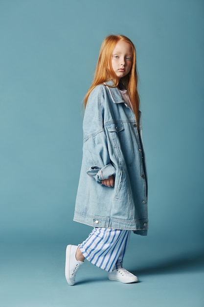 Bella bambina dai capelli rossi con i capelli lunghi in una grande giacca di jeans blu lungo.