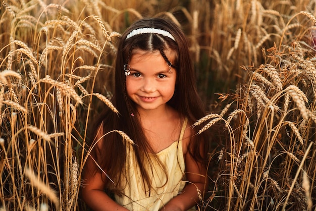 Bella bambina di aspetto europeo in piedi in un campo di grano in un vestito giallo con un sorriso sul viso al tramonto Felice dolce bambina
