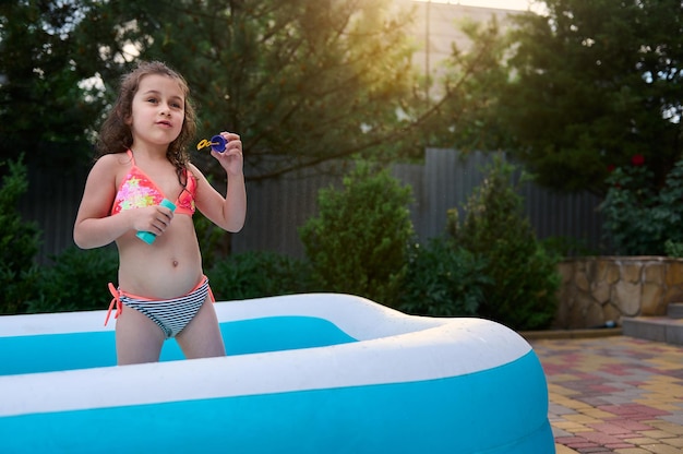 Bella bambina europea in costume da bagno soffia palline di sapone divertendosi in una piscina d'acqua gonfiabile nel cortile di casa