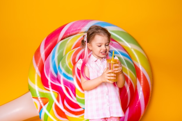 Bella bambina felice in vestito rosa che beve succo d'arancia su superficie gialla con lecca-lecca.