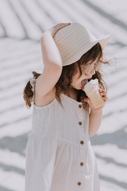 Bella bambina in occhiali da sole vestito bianco e cappello che mangia il gelato sulla spiaggia in estate