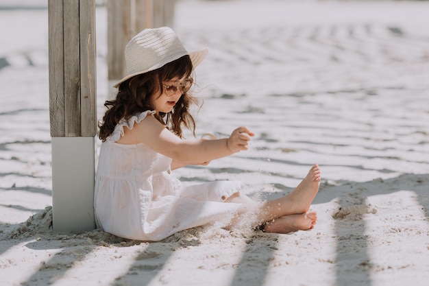 Bella bambina in occhiali da sole vestito bianco e cappello che mangia il gelato sulla spiaggia in estate