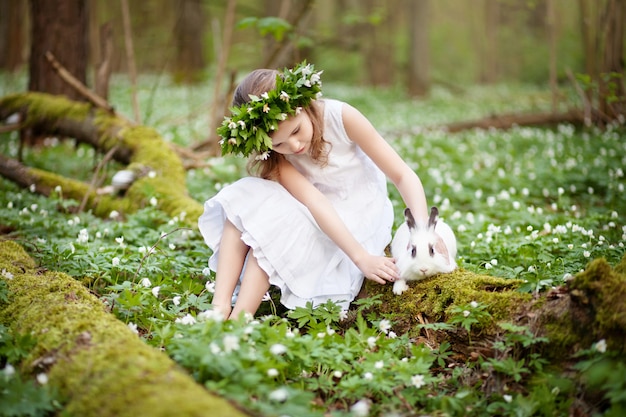 Bella bambina in un abito bianco che gioca con coniglio bianco nel legno di primavera.