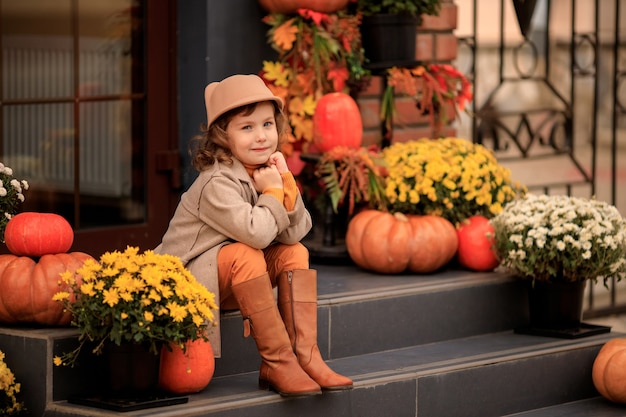 Bella bambina in un cappello sui gradini della casa con zucche e fiori