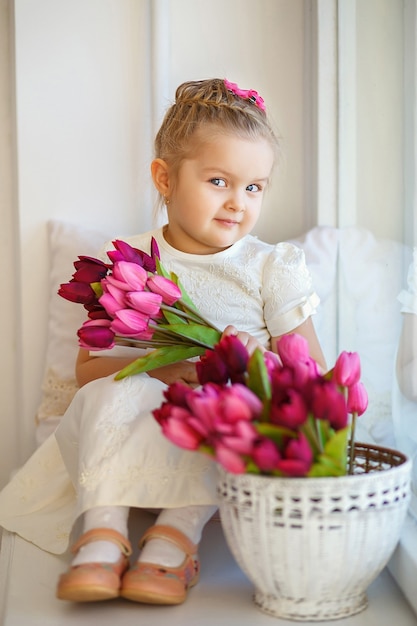 Bella bambina in un vestito bianco che si siede su un davanzale con un mazzo di tulipani rosa