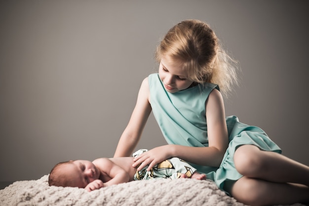 Bella bambina in un vestito blu guarda il suo piccolo affascinante fratello neonato addormentato sdraiato su un plaid su uno sfondo grigio