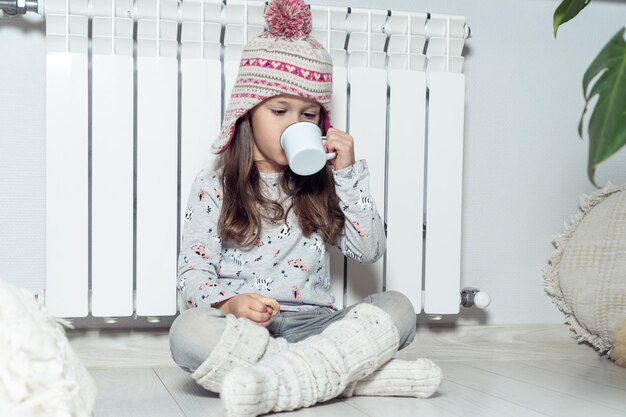 Bella bambina riccia in calze di lana cappello lavorato a maglia mangiare dolce dessert bere cacao caldo sedersi vicino al radiatore di riscaldamento bianco