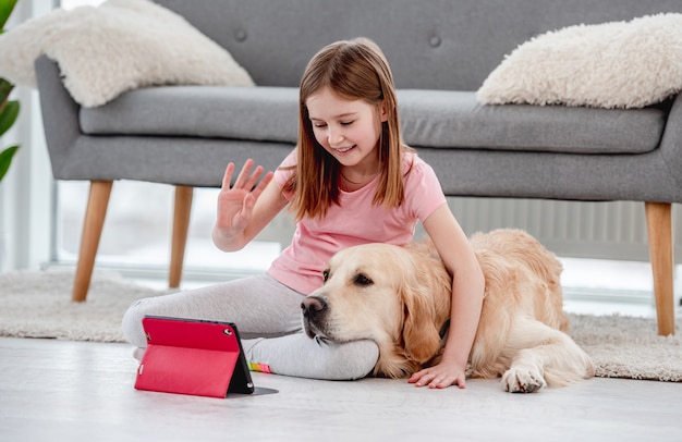 Bella bambina seduta sul pavimento con il cane golden retriever durante la comunicazione online con gli amici sul tablet