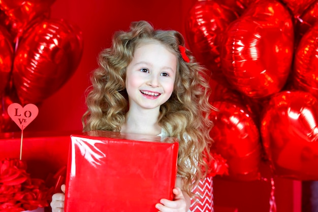 Bella bambina sorride su uno sfondo rosso con un palloncino a forma di cuore e una confezione regalo in mano Il concetto di regali d'amore San Valentino 14 febbraio 8 marzo festa della mamma