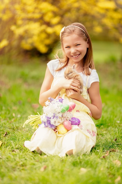 Bella bambina sorridente che tiene coniglietto carino e bouquet di fiori bianchi e uova di Pasqua nelle vacanze primaverili.