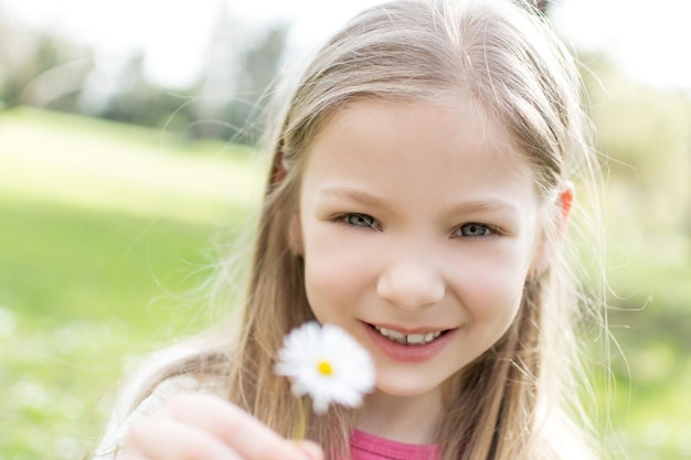 Bella bambina sorridente con gli occhi azzurri nel parco che tiene fiore bianco e che guarda l'obbiettivo.