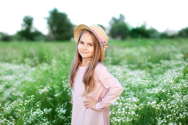 Bella bambina sorridente in abito rosa e cappello di paglia in campo di margherite. Bambino sveglio nel campo della camomilla di fioritura in estate. Fiori selvatici.